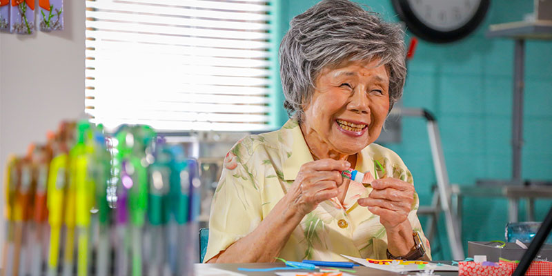 A smiling elderly woman with short grey hair works on a small craft at a table.