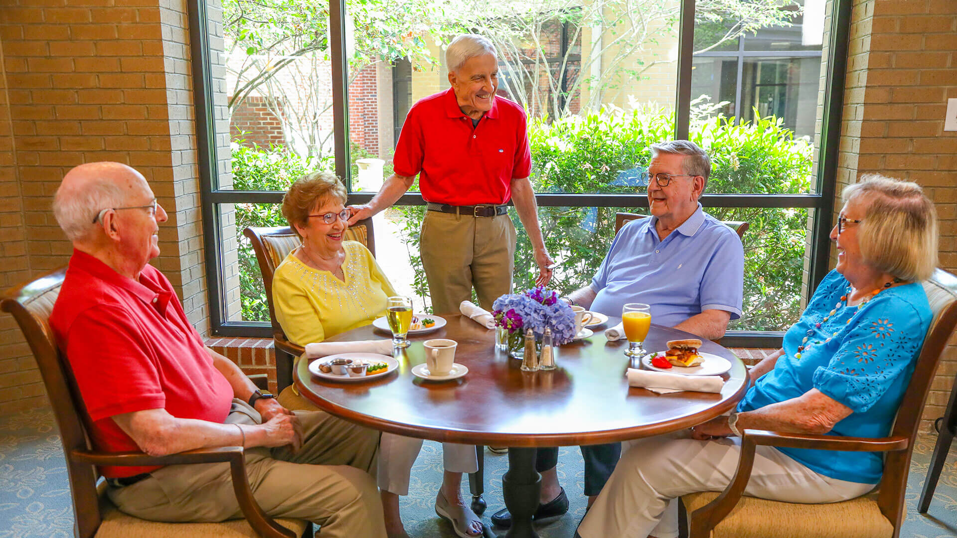 Five seniors smiling and talking around a dining table near a large window.
