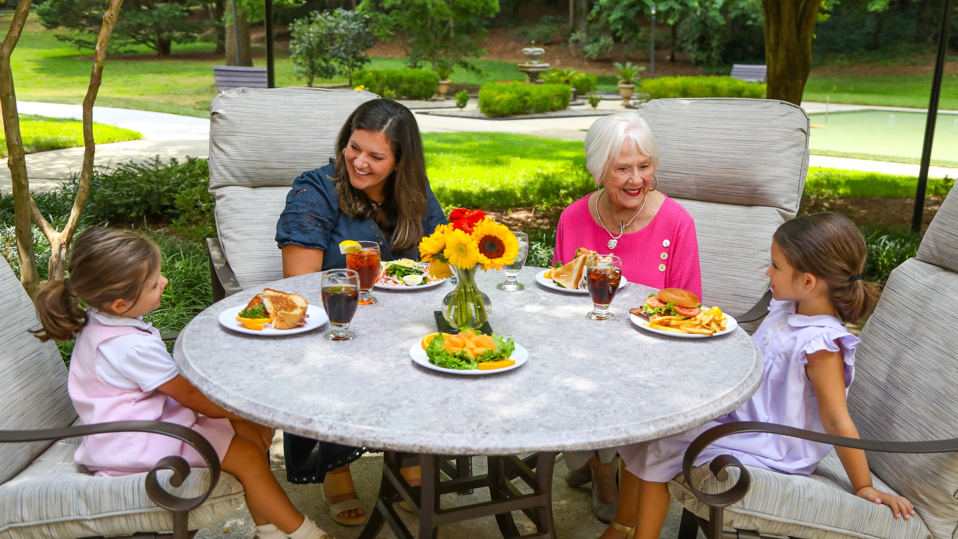 A multi-generational family enjoys lunch together at a round patio table in a lush outdoor garden.
