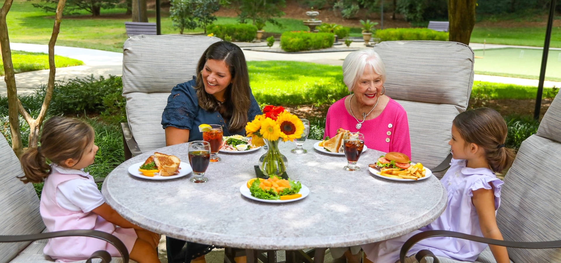 A multi-generational group enjoys a meal together at an outdoor patio table surrounded by lush greenery.