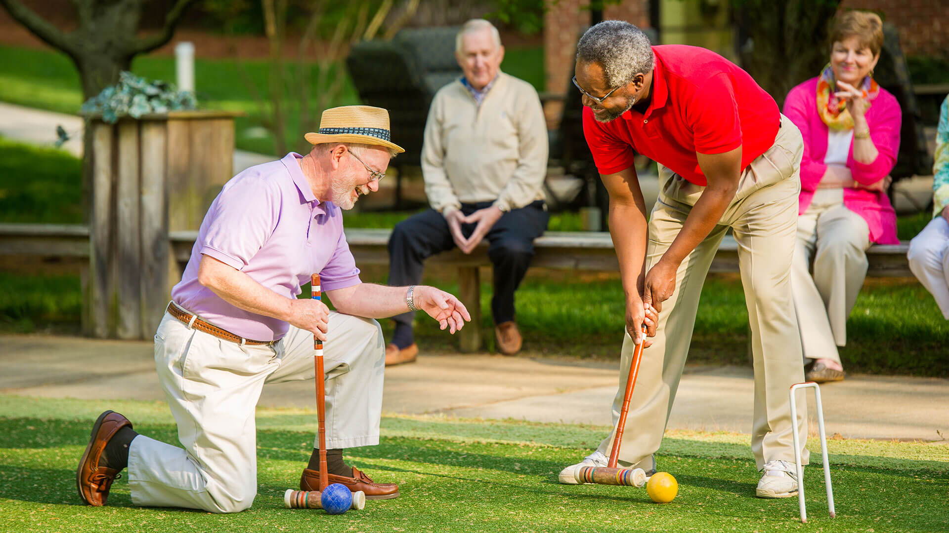 Two older men play croquet on a lawn while friends watch from a bench in the background.