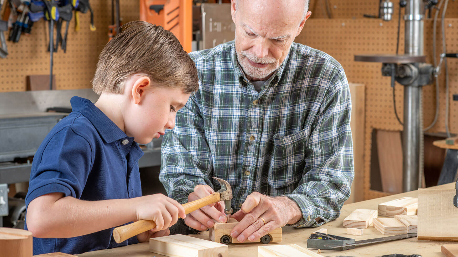 An older man helps a young boy hammer a nail into a wooden toy car in a workshop.