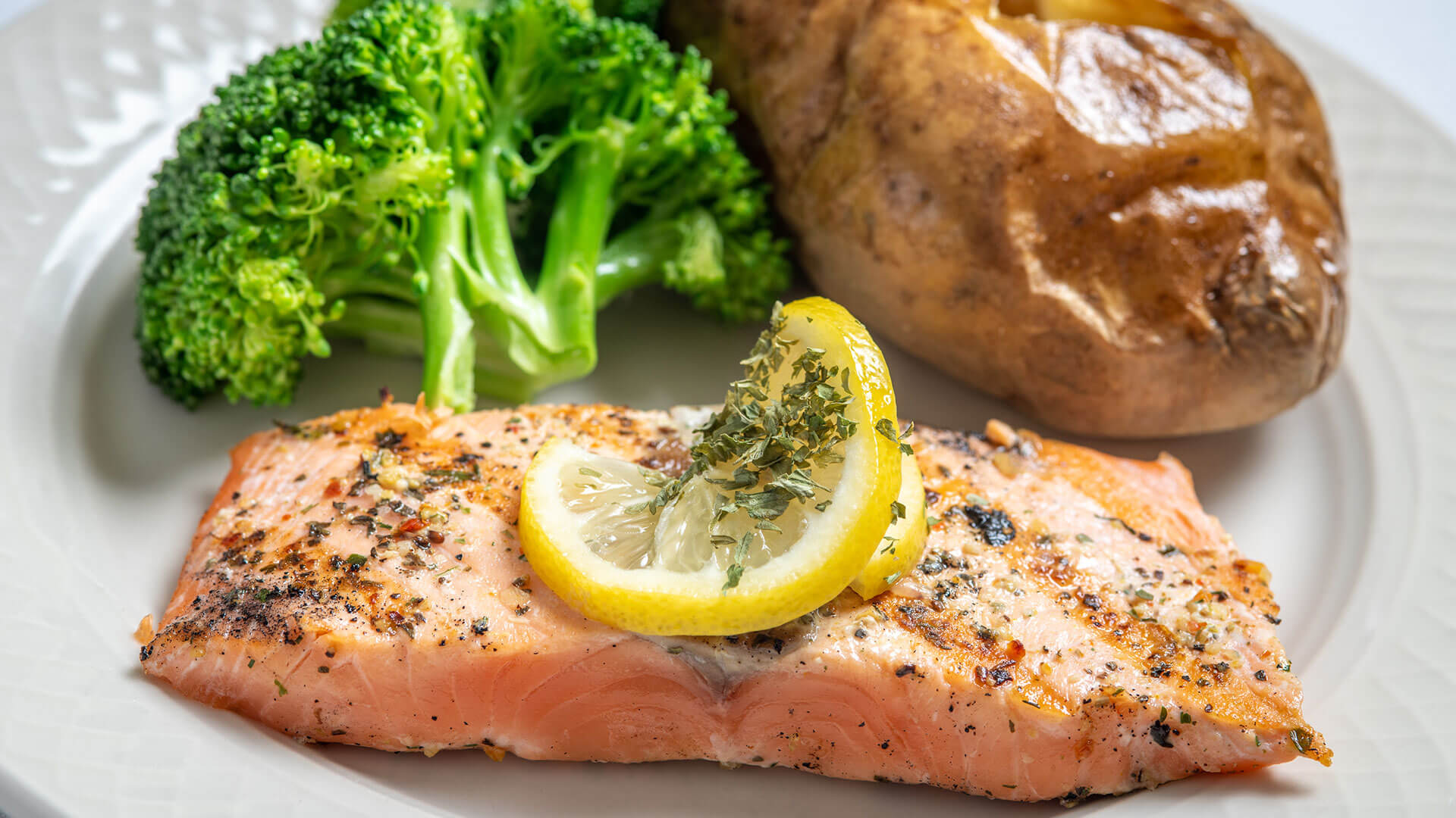 Seasoned salmon with a lemon slice, steamed broccoli, and a baked potato on a white plate.