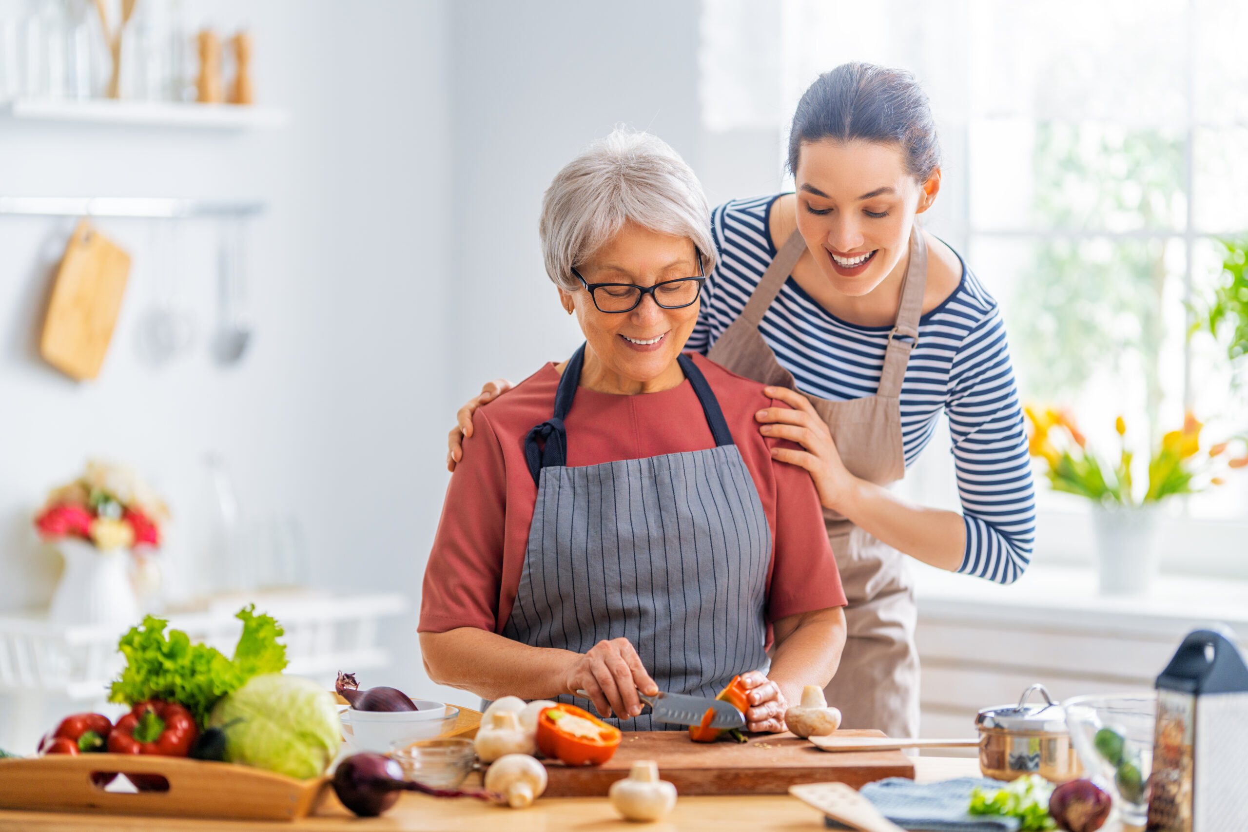 A younger woman smiling with her hands on an older woman's shoulders as they prepare vegetables in a kitchen.