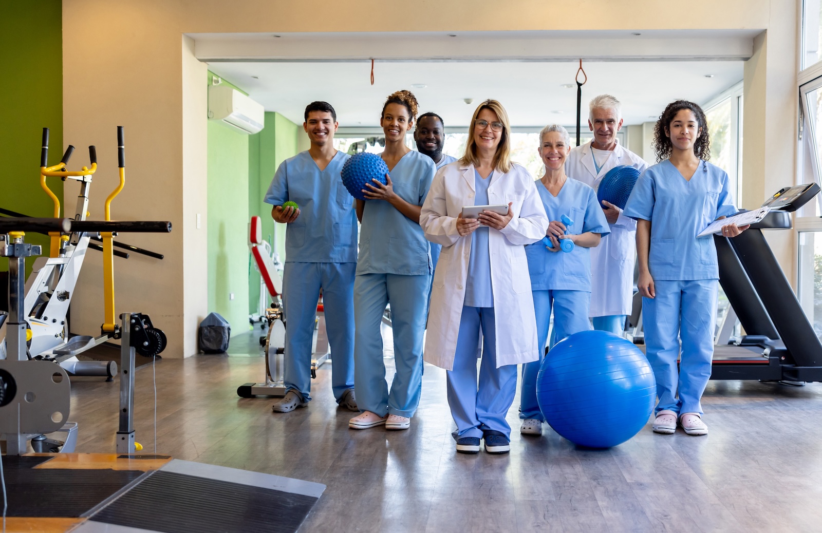 A smiling team of healthcare professionals in blue scrubs and a white lab coat standing in a physical therapy gym.