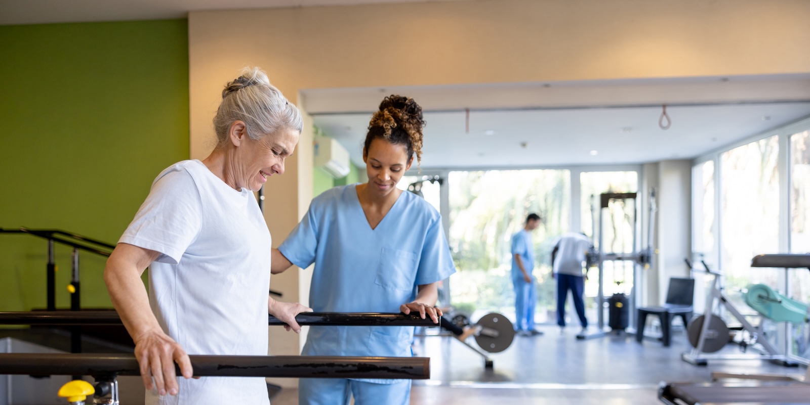 A physical therapist assists an elderly woman walking between parallel bars in a bright gym.