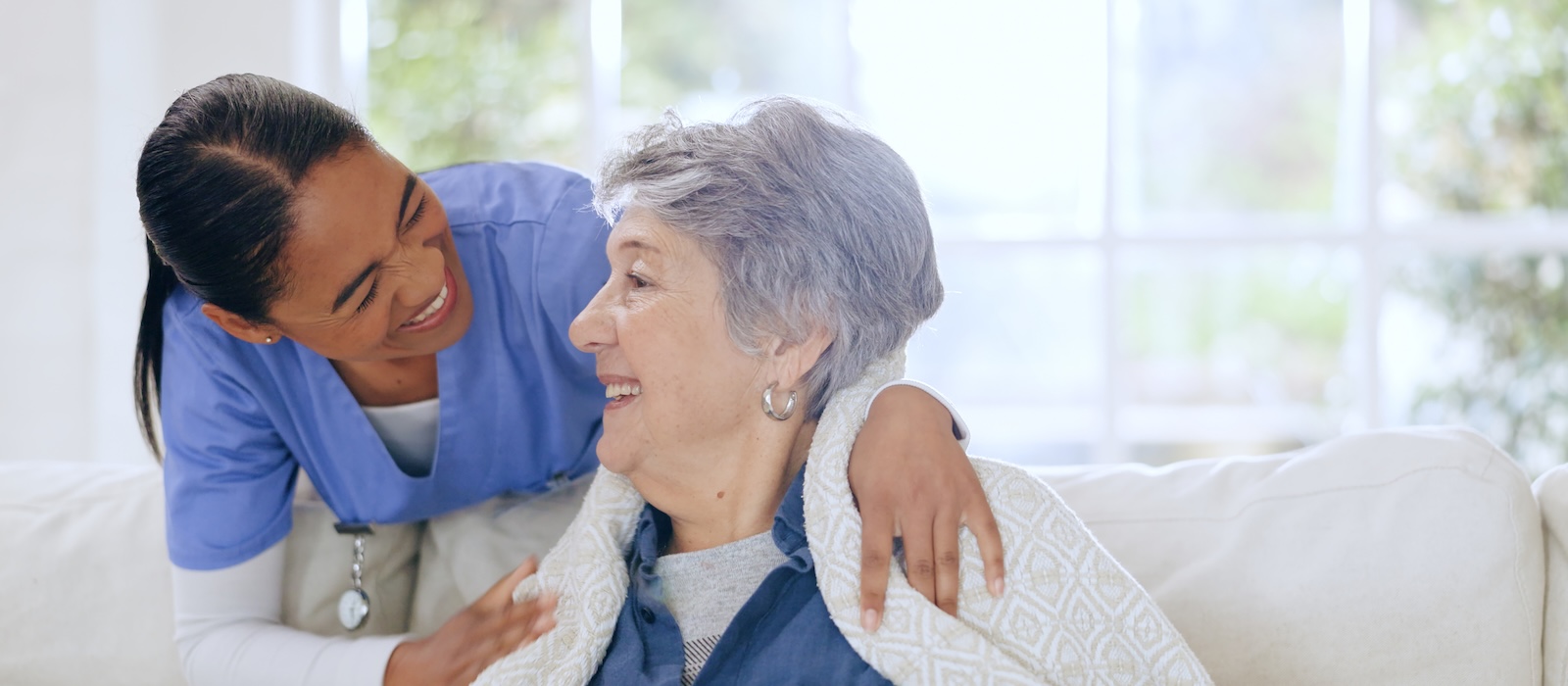 A smiling caregiver in blue scrubs embraces an elderly woman wrapped in a blanket.
