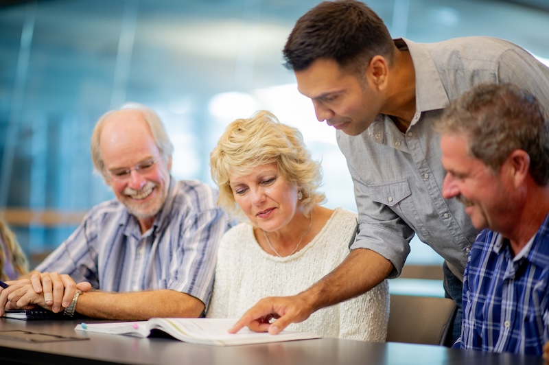 A man points at a book for three smiling senior adults sitting at a table.